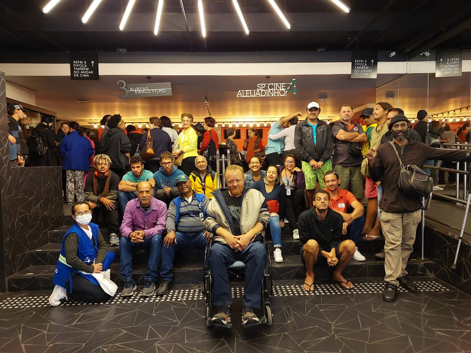 A foto mostra um grupo com cerca de 20 pessoas sentadas no hall de um cinema; acima deles, vê-se a indicação de salas de cinema; em primeiro plano, entre as pessoas que estão posando para a foto, ,está um homem sentado em uma cadeira de rodas